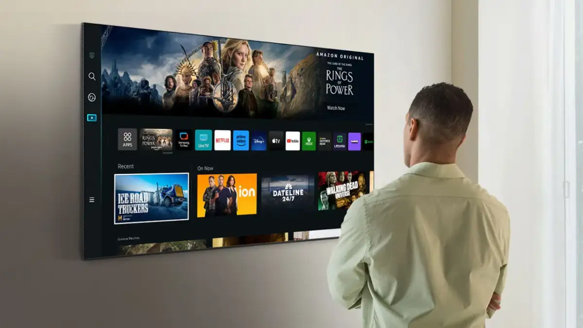 Mężczyzna patrzy na ekran telewizora Samsung Smart TV, gdzie wyświetlane są aplikacje i filmy. Dowiedz się, jak zaktualizować telewizor Samsung Smart TV, aby cieszyć się najnowszymi funkcjami.