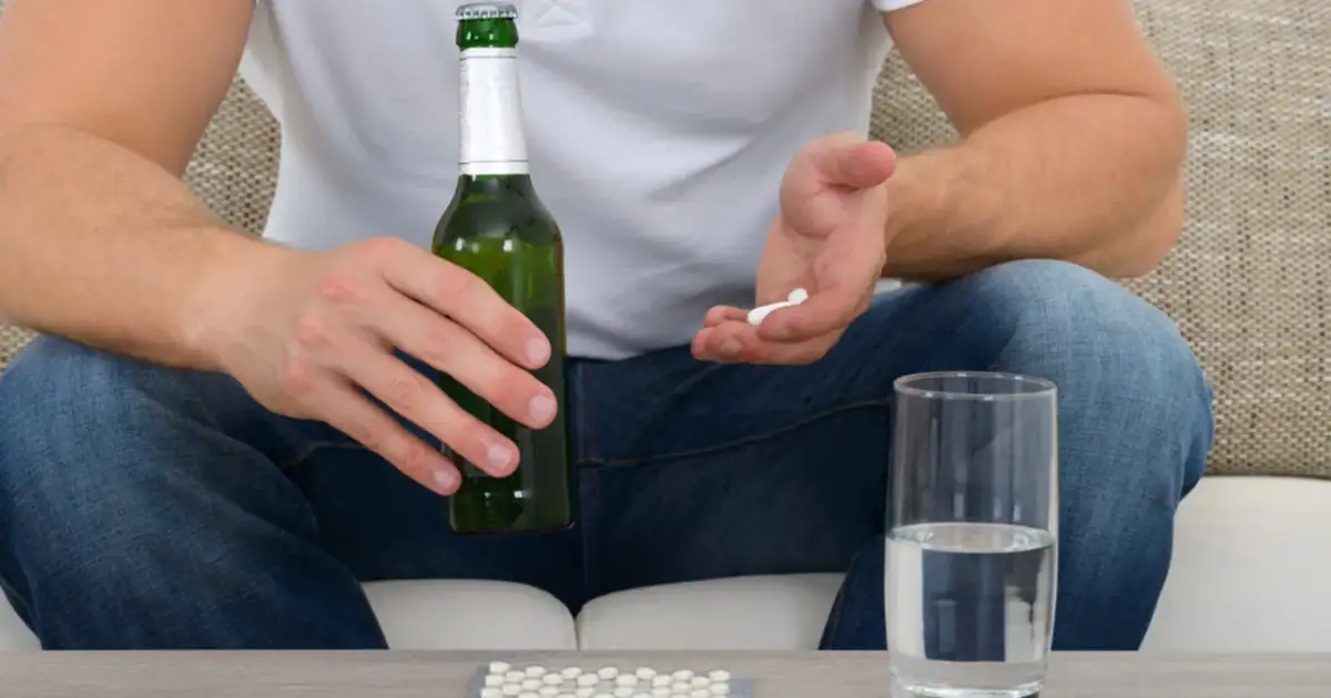 Mężczyzna trzyma butelkę piwa i garść tabletek, obok stoi szklanka wody. Czy to ibuprofen i alkohol?