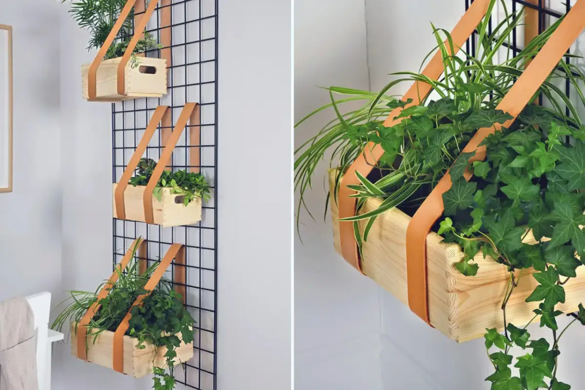 Prosty sposób, jak zrobić kwietnik na ścianę: drewniane skrzynki z roślinami wiszą na czarnej siatce, trzymane przez skórzane paski.