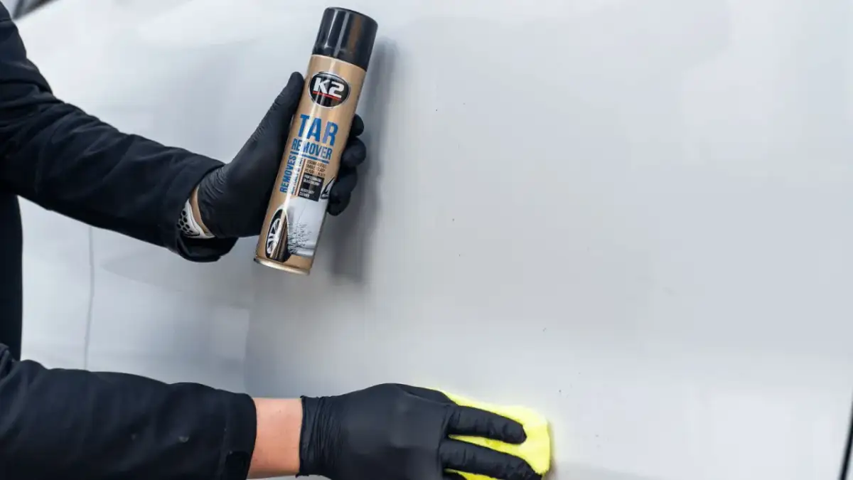 Ręce w czarnych rękawiczkach używają preparatu K2 Tar Remover, by usunąć naklejki z samochodu. Żółta ściereczka poleruje powierzchnię.