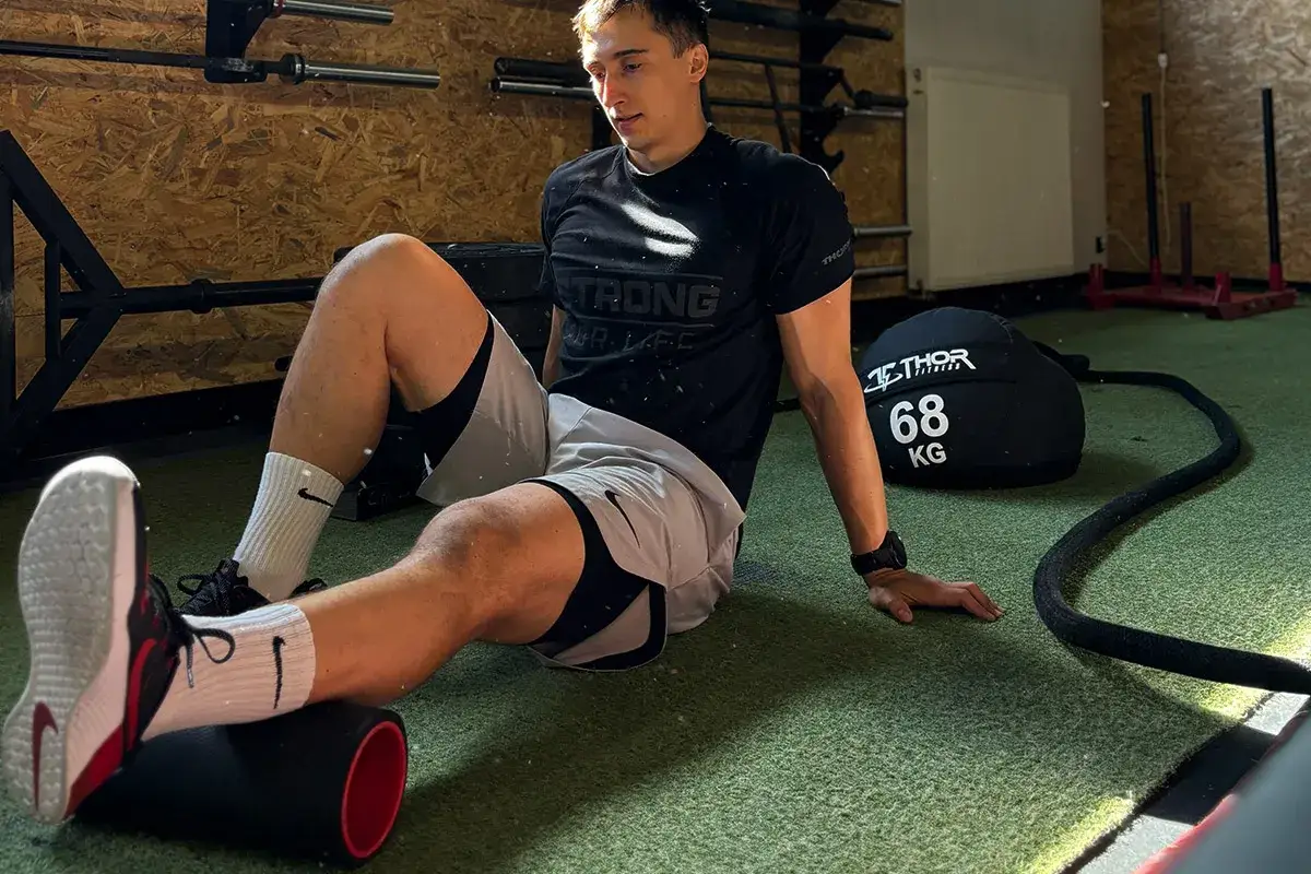 Mężczyzna na siłowni roluje mięśnie na wałku. To część treningu, który może być związany z metcon crossfit, co to znaczy intensywny wysiłek.