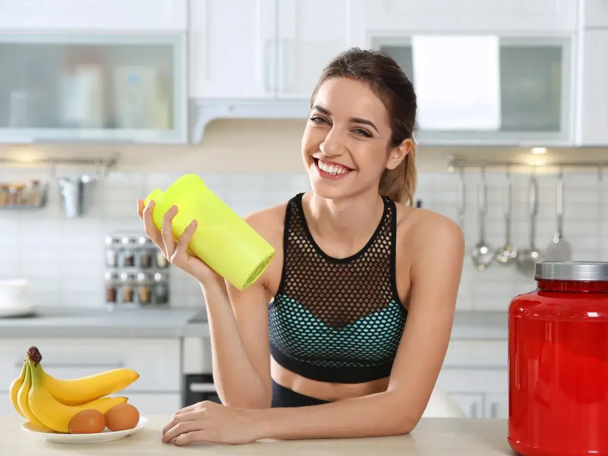 Uśmiechnięta kobieta z żółtym shakerem w ręku, obok banany, jajka i duży czerwony pojemnik z odżywką. Zastanawia się, czy brać kreatynę w dni bez treningu.