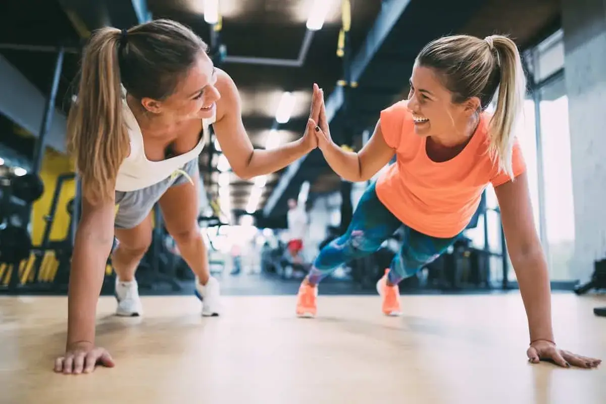 Dwie uśmiechnięte kobiety robią pompki w siłowni, świętując sukces. Chcesz wiedzieć, jak zdobyć kartę Multisport?