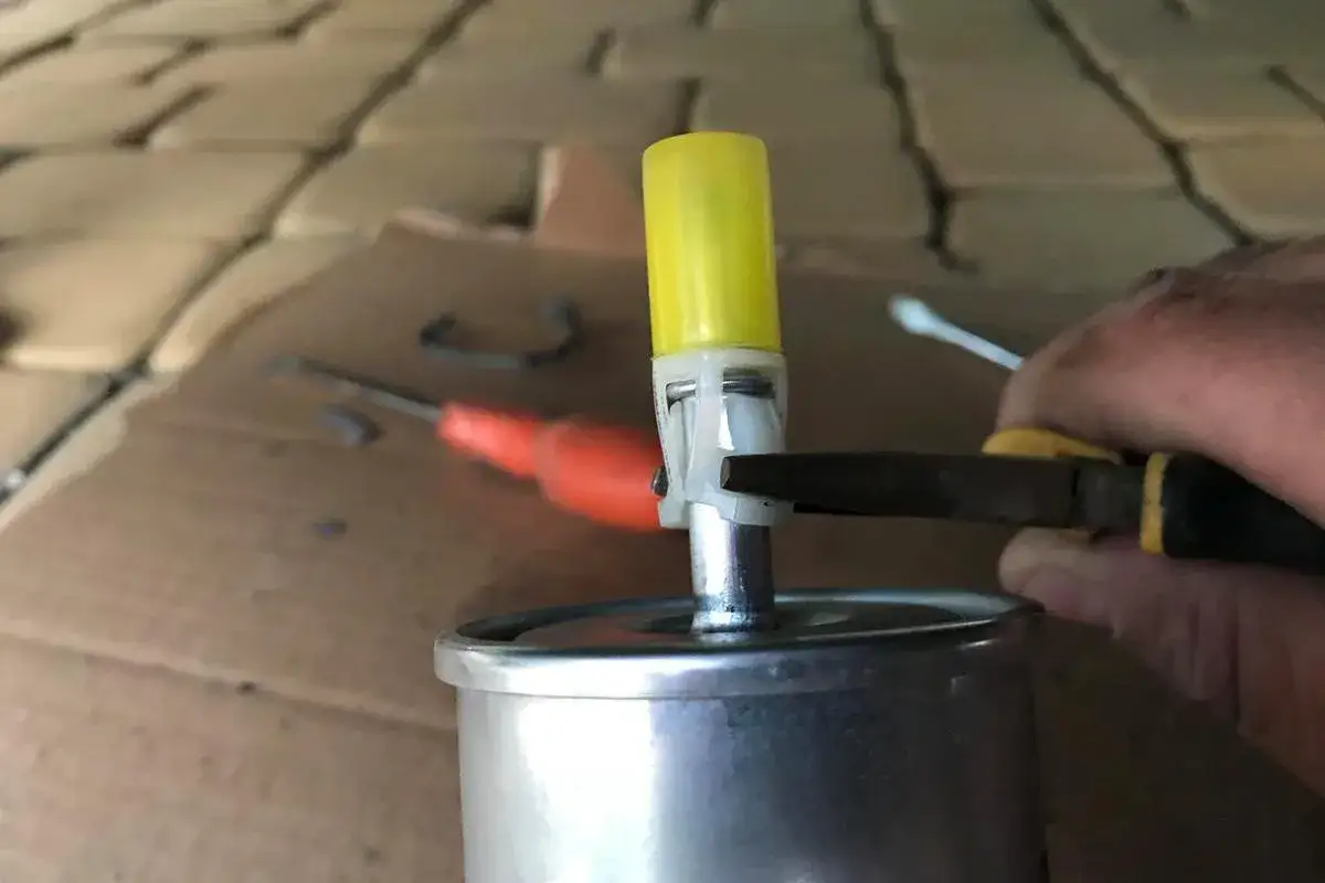 Dłoń trzymająca szczypce przy filtrze paliwa z żółtą zatyczką.