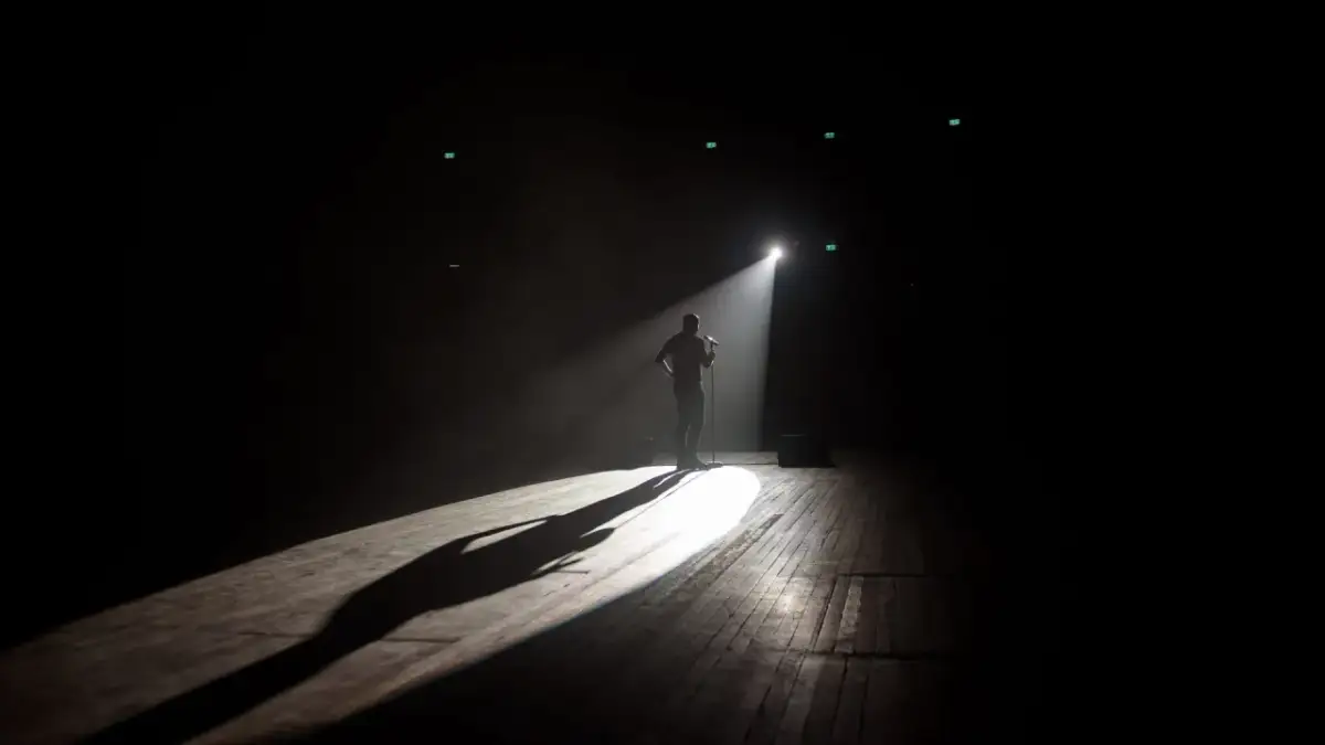 Mężczyzna stoi na scenie w świetle reflektora, gotowy do występu na open mike stand up. Długi cień rzuca się na drewnianą podłogę.