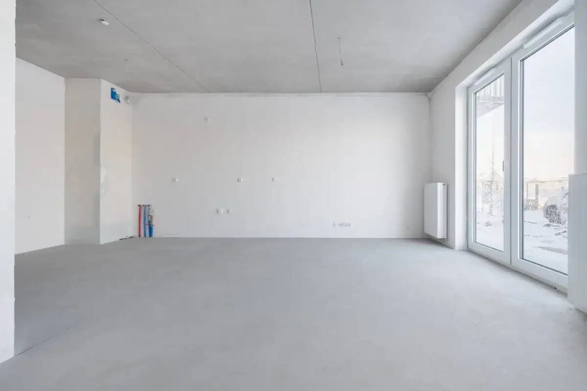 Pusta przestrzeń w stanie deweloperskim: betonowa posadzka, białe ściany, instalacje wodne i elektryczne, grzejnik, duże okna.