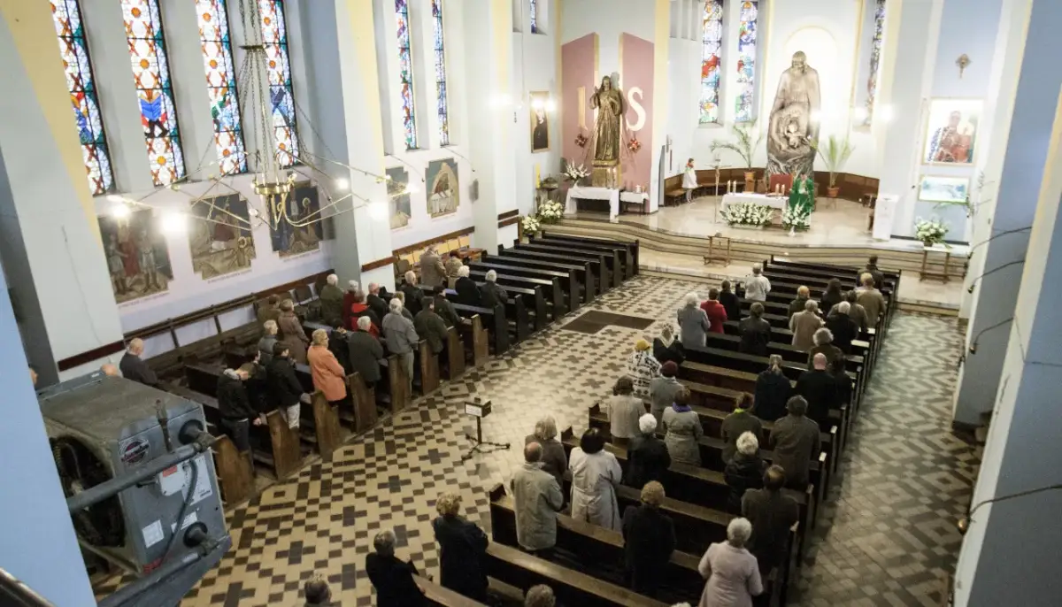 Ludzie siedzą w ławkach podczas mszy wieczornej przed świętem. Witraże i obrazy zdobią wnętrze kościoła.