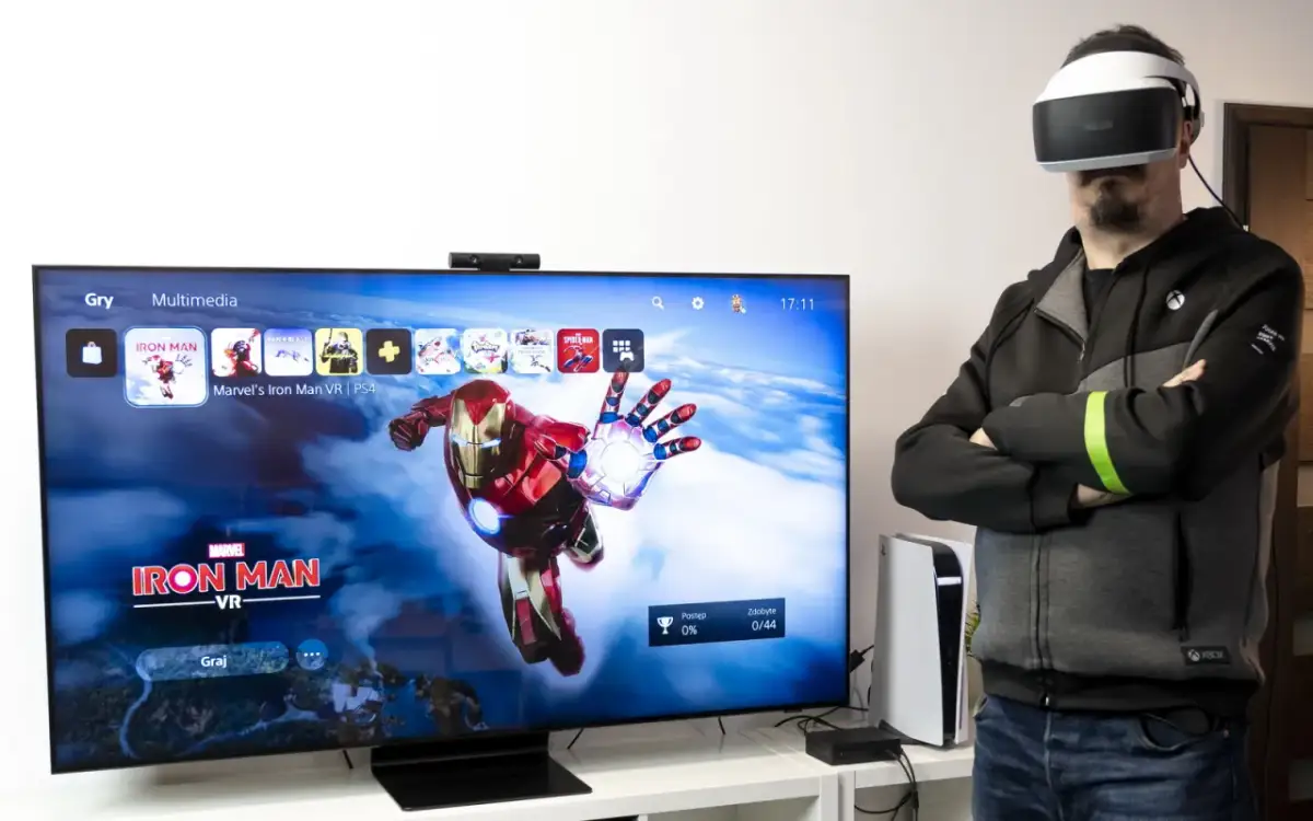 Mężczyzna w goglach VR obok telewizora z grą Iron Man VR.