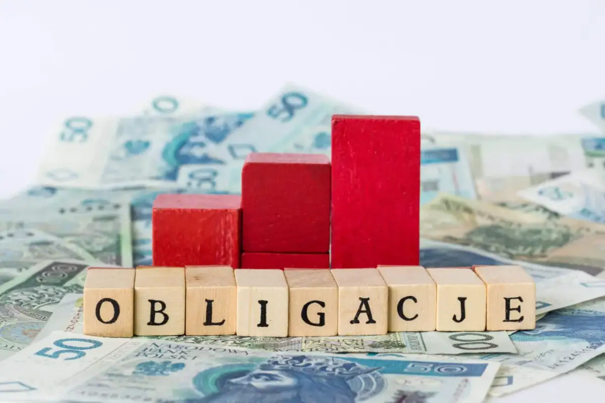 Drewniane klocki z napisem "OBLIGACJE" na tle polskich banknotów. Symbolizują one, kto emituje obligacje skarbowe.