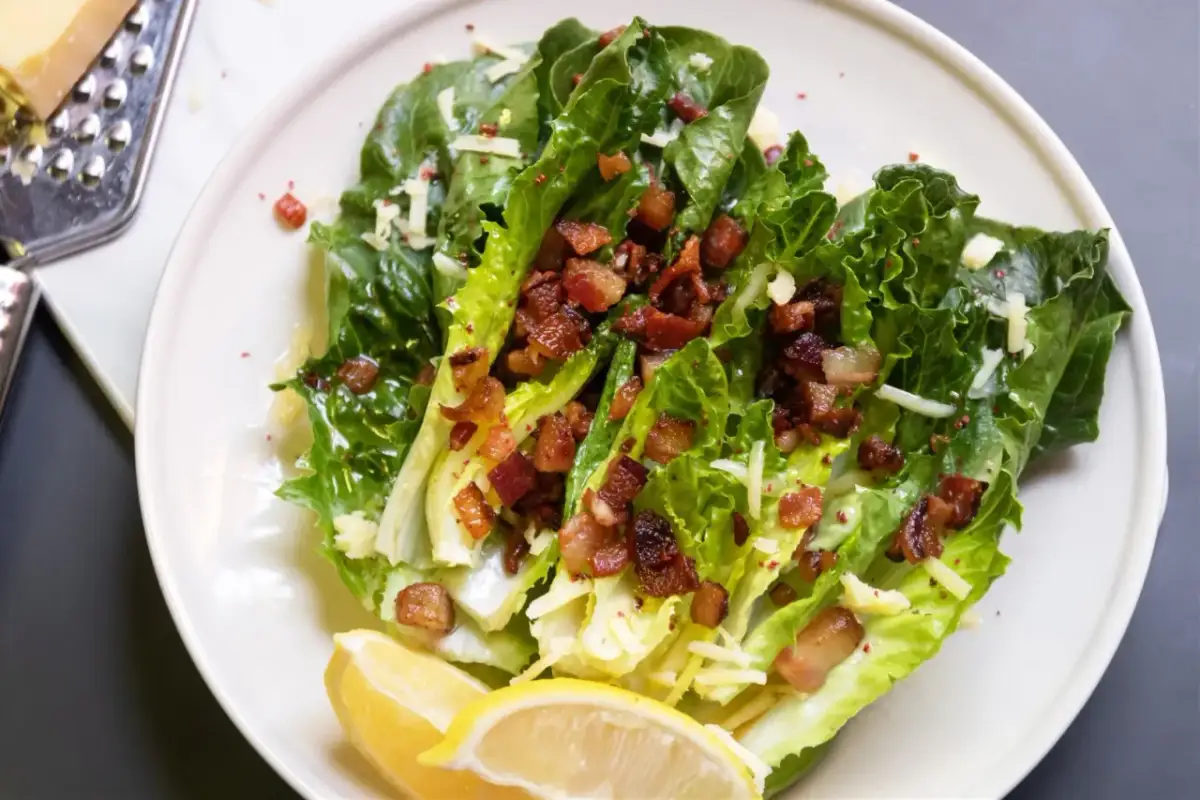 Sałatka cezar z boczkiem, chrupiące liście sałaty rzymskiej, posypane startym parmezanem i kawałkami chrupiącego boczku. Podana z cytryną.
