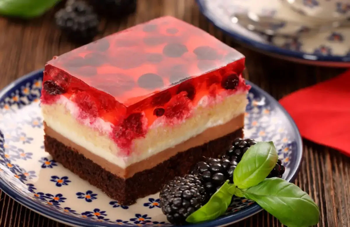 Kawałek ciasta z galaretką owocową, kremem i biszkoptem na talerzyku z jeżynami.