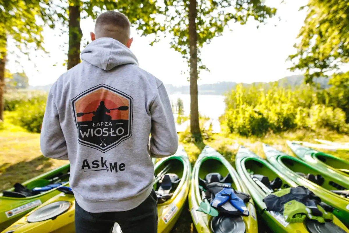 Gotowy na spływy kajakowe? Na zdjęciu mężczyzna w bluzie z logo "Łap za wiosło" stoi obok rzędu kajaków nad jeziorem.