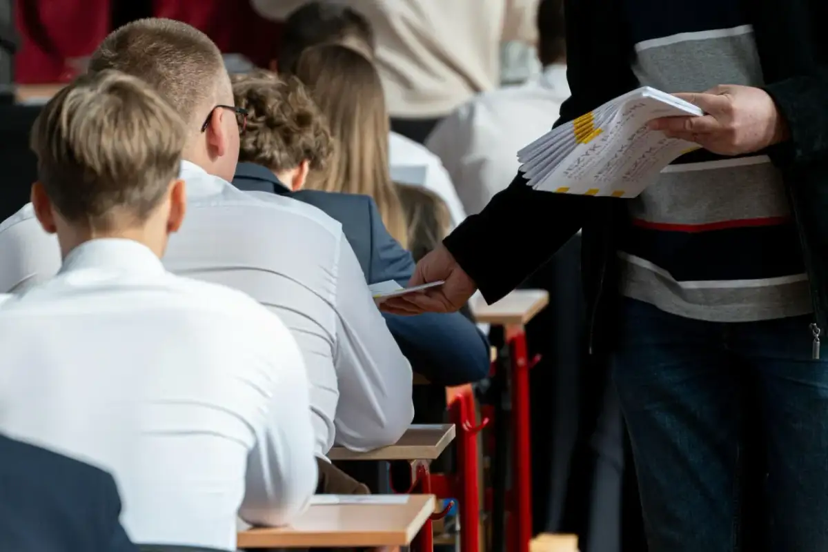 Nauczyciel rozdaje arkusze egzaminacyjne uczniom siedzącym w ławkach.
