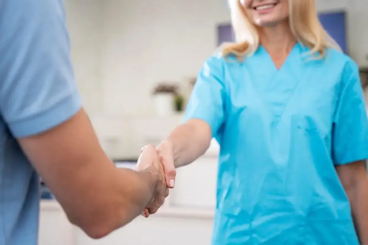 Uśmiechnięta opiekunka w niebieskim fartuchu wita pacjenta uściskiem dłoni. Kiedy należy się opiekunka z mops?