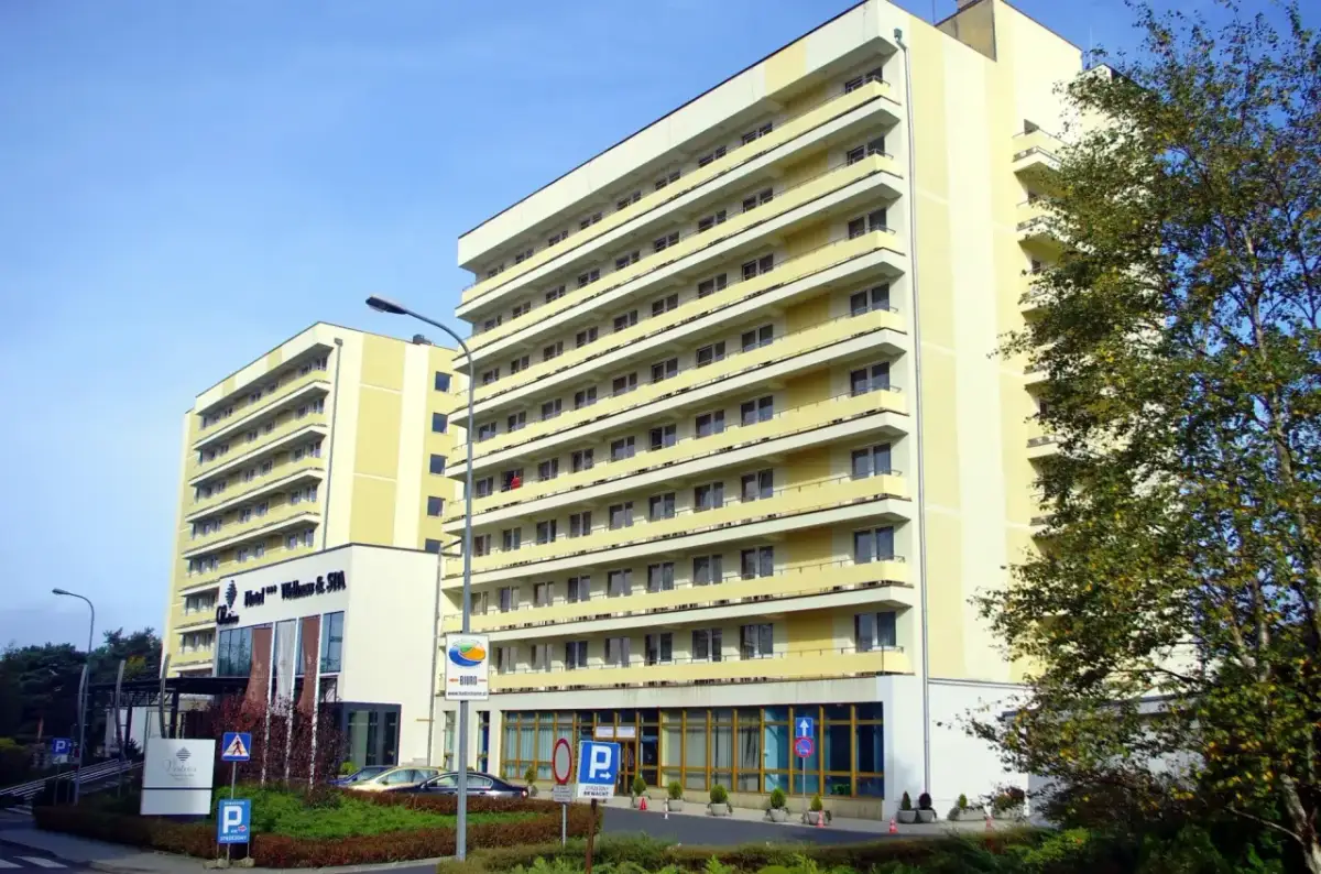 Hotel Vestina w Międzyzdrojach, wysoki, żółty budynek z balkonami, otoczony zielenią.