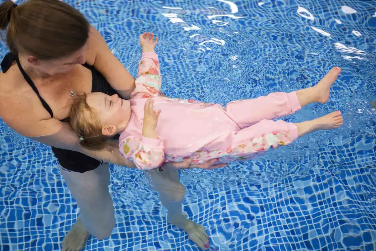 Dziecko w różowym kombinezonie do pływania dla dzieci unosi się na wodzie, trzymane przez instruktorkę.