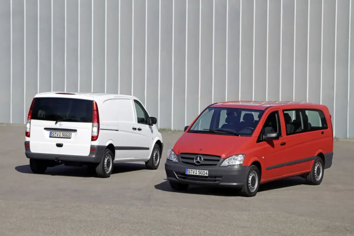 Dwa busy Mercedes-Benz: biały towarowy i czerwony osobowy, stojące na tle szarej ściany.