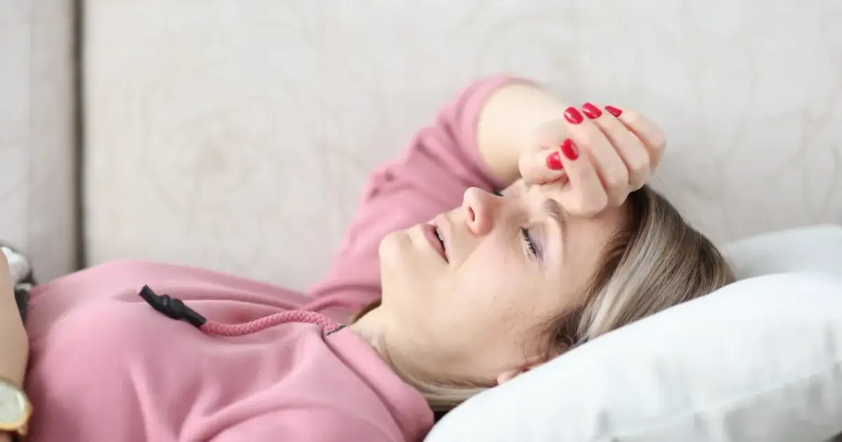 Kobieta w różowej bluzie leży na kanapie z ręką na czole, z zamkniętymi oczami.