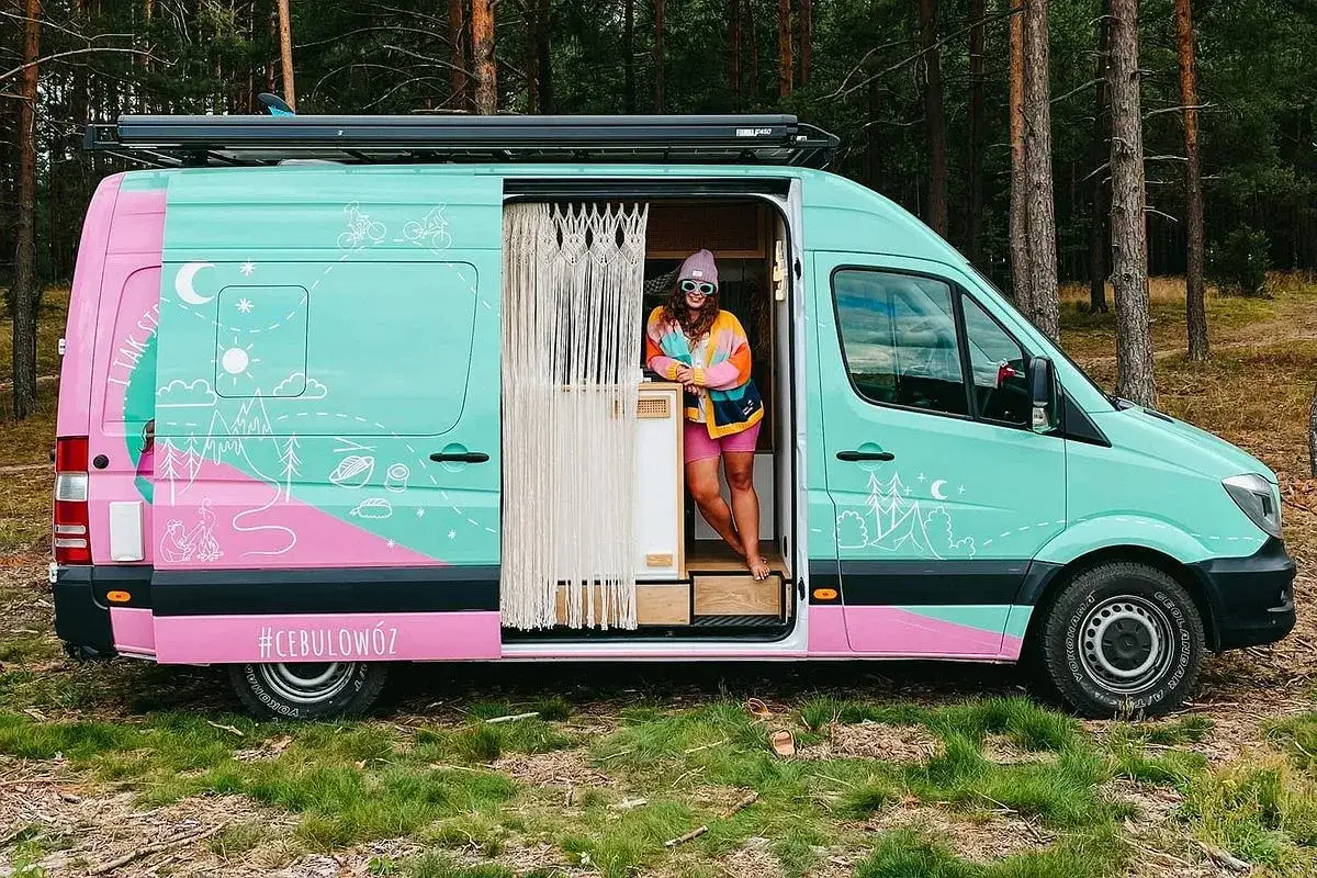 Kolorowy minivan camper z rysunkami przyrody i napisem #CEBULOWÓZ. Kobieta w kolorowym swetrze stoi w otwartych drzwiach.