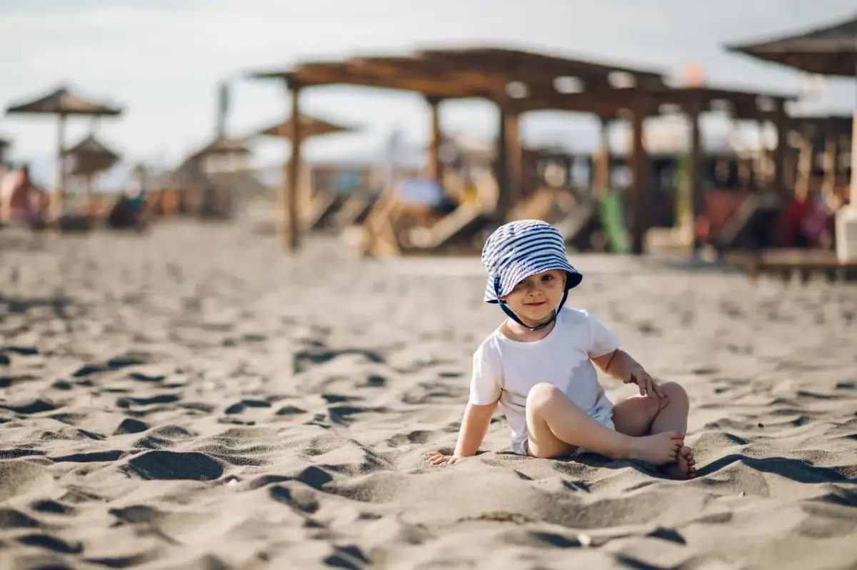 Małe dziecko w pasiastym kapeluszu siedzi na piaszczystej plaży, w tle widać parasole i leżaki.