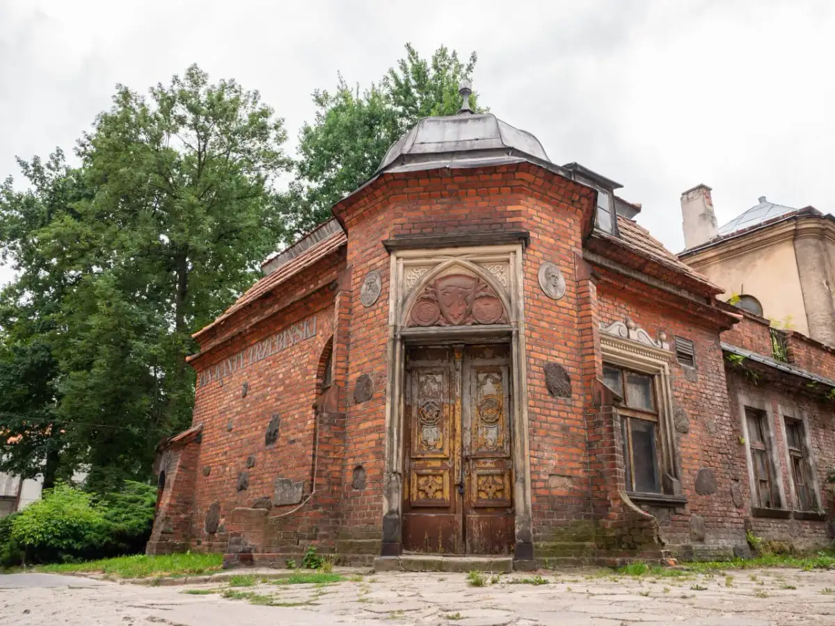 Stary ceglany budynek z ozdobnymi drzwiami i medalionami, otoczony zielenią.