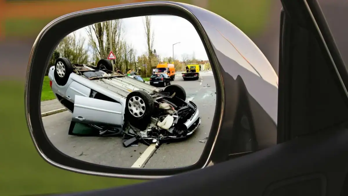 Auto w leasingu leży na dachu po wypadku. Widok w lusterku bocznym pokazuje szkody z OC sprawcy i pomoc drogową na drodze.