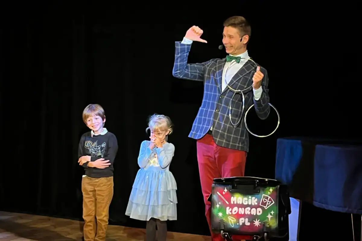 Magik Konrad wyjaśnia dzieciom, co to jest teatr, podczas pokazu iluzji. Dzieci z zaciekawieniem obserwują sztuczki.