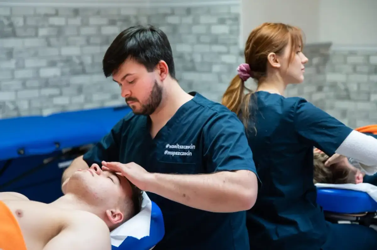 Fizjoterapeuta wykonuje masaż twarzy pacjentowi, w tle druga fizjoterapeutka.