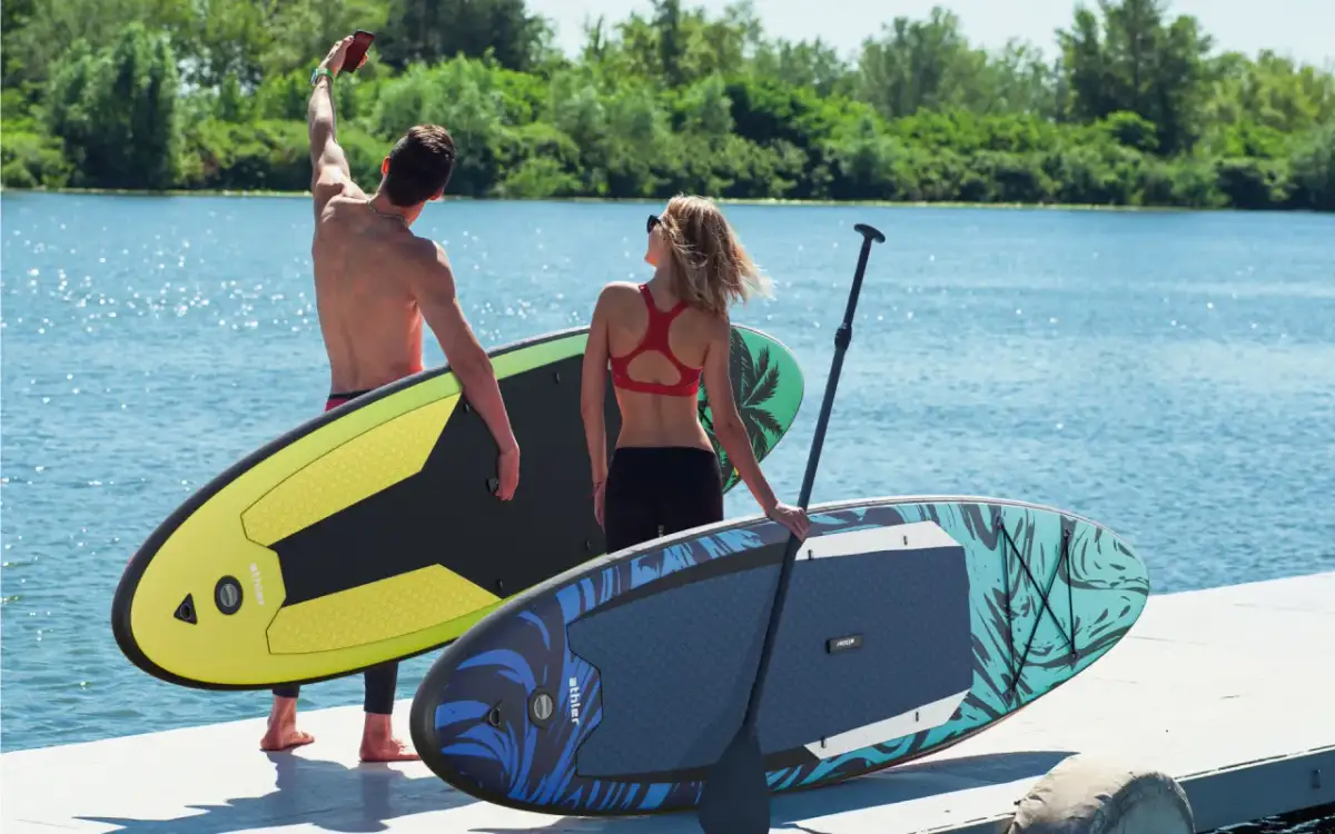 Para szykuje się na wodne przygody. Mężczyzna robi selfie, trzymając w ręku telefon, a obok niego stoi kolorowa deska do pływania.