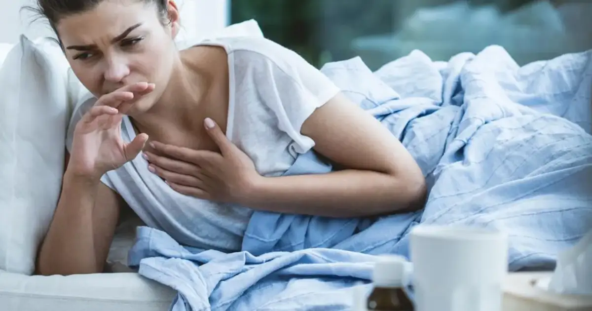 Kobieta w łóżku kaszle, zakrywając usta dłonią. Czuje się źle, może ma brązową flegmę.