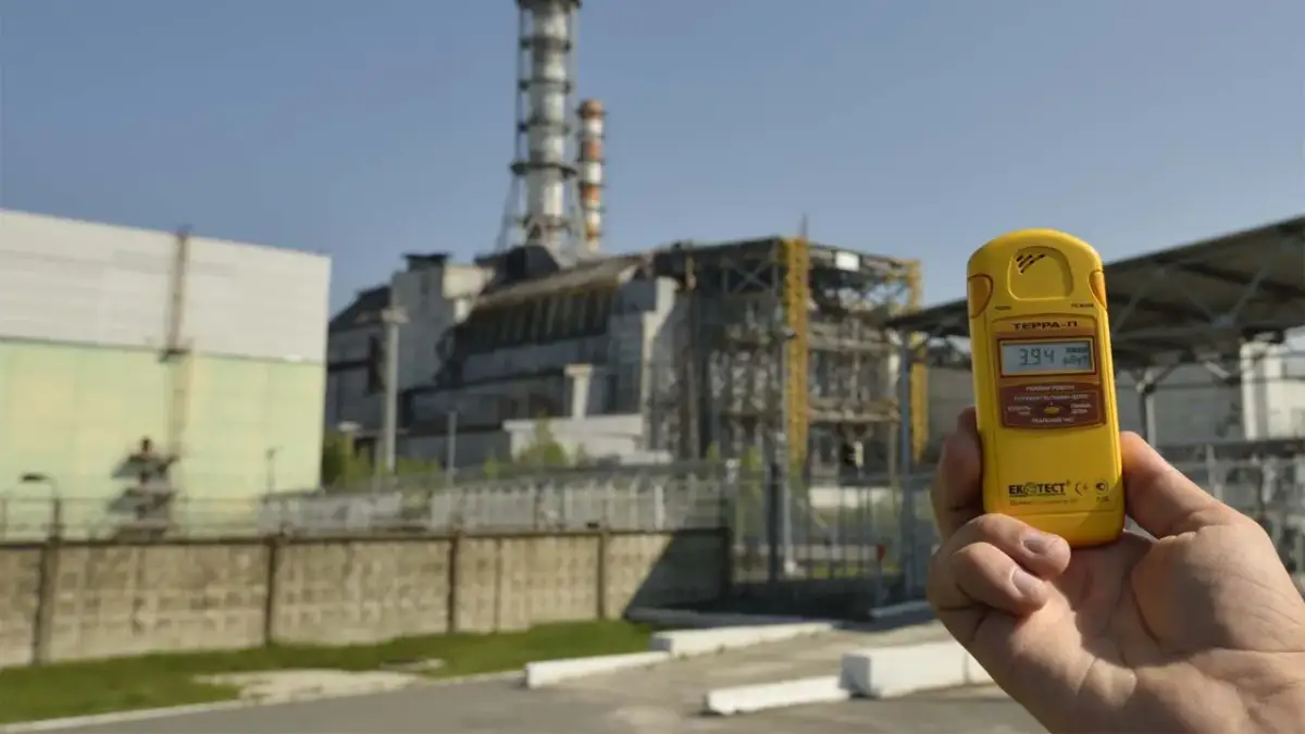 Żółty dozymetr pokazuje 3.94 µSv/h przed budynkami elektrowni w Czarnobylu, odpowiadając na pytanie, czy Czarnobyl dalej promieniuje.