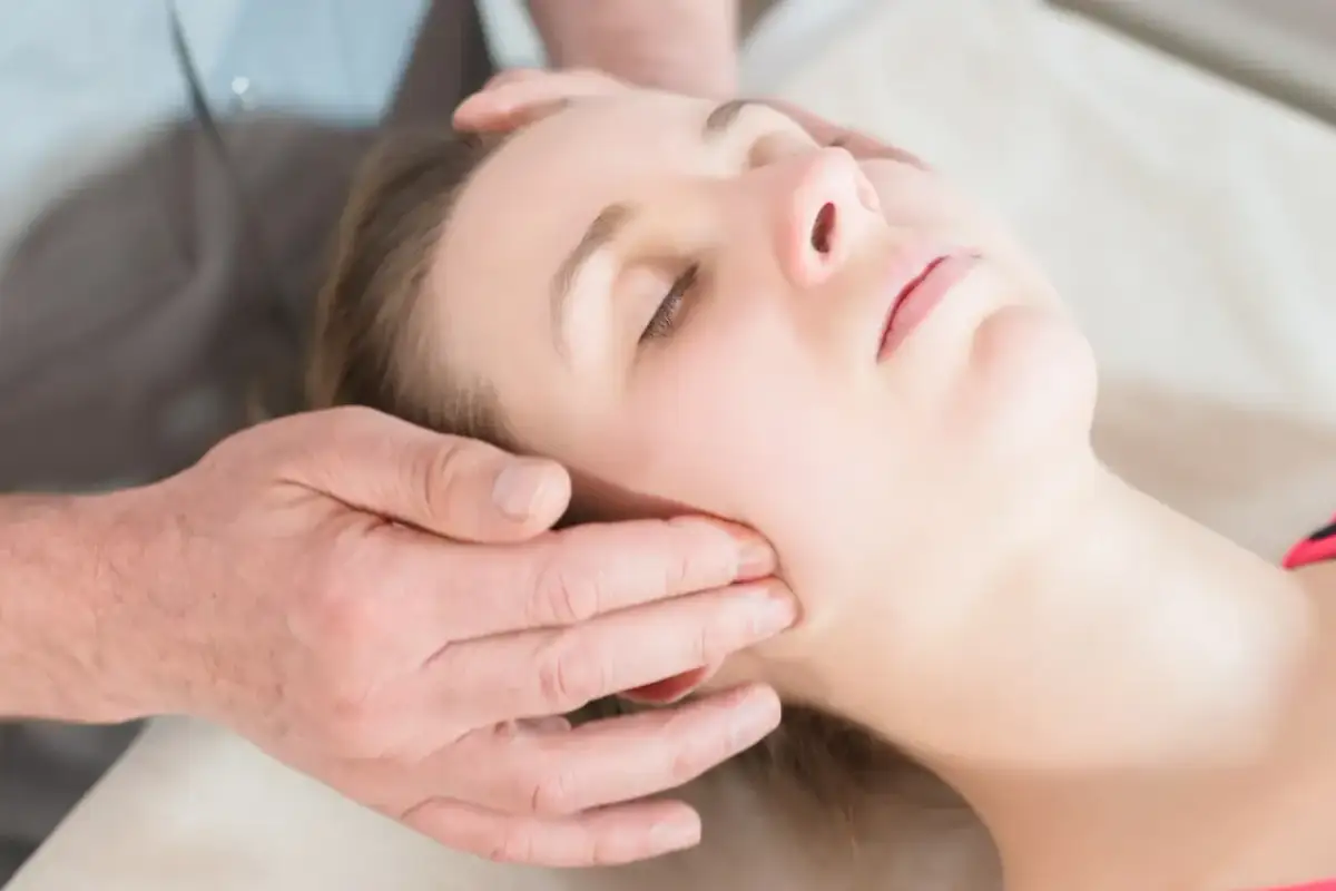 Delikatny masaż twarzy, który pomaga jak rozluźnić szczękę. Dłonie terapeuty obejmują policzek i skroń.