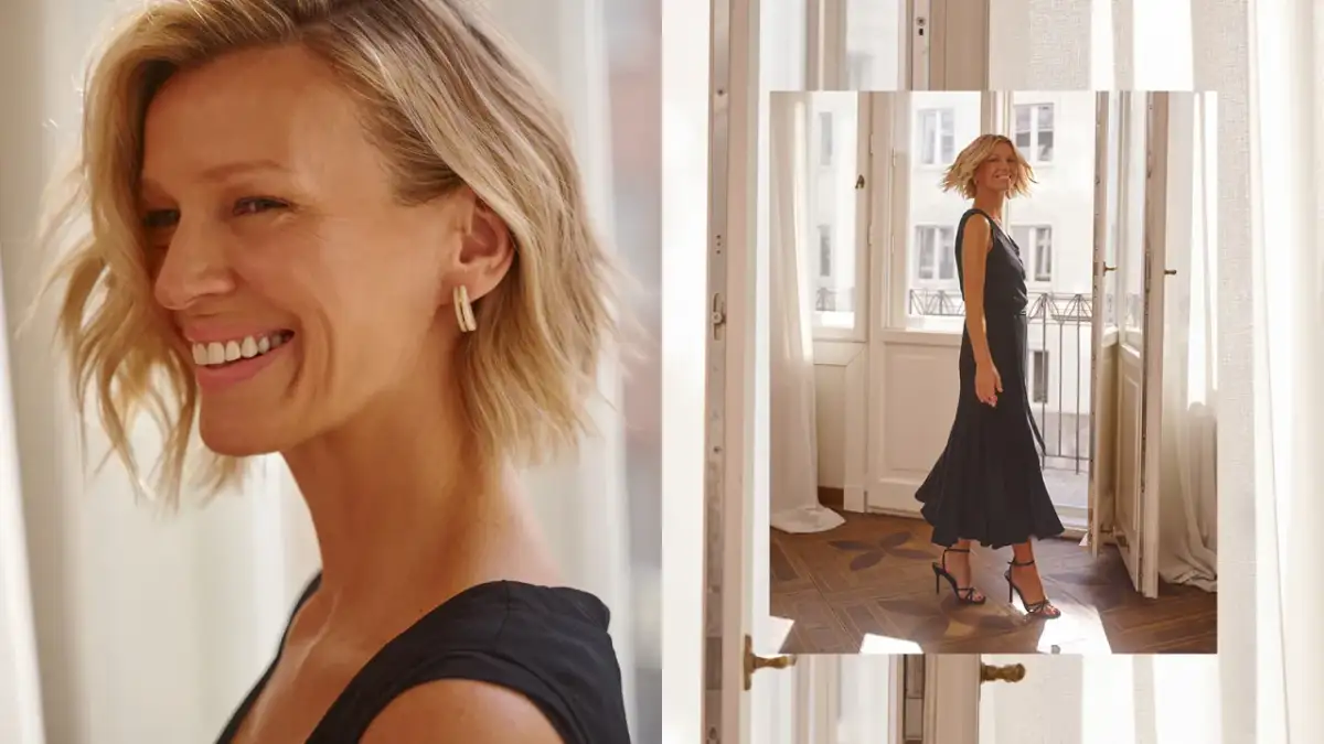 Uśmiechnięta kobieta w czarnej sukience i szpilkach stoi przy drzwiach balkonowych.