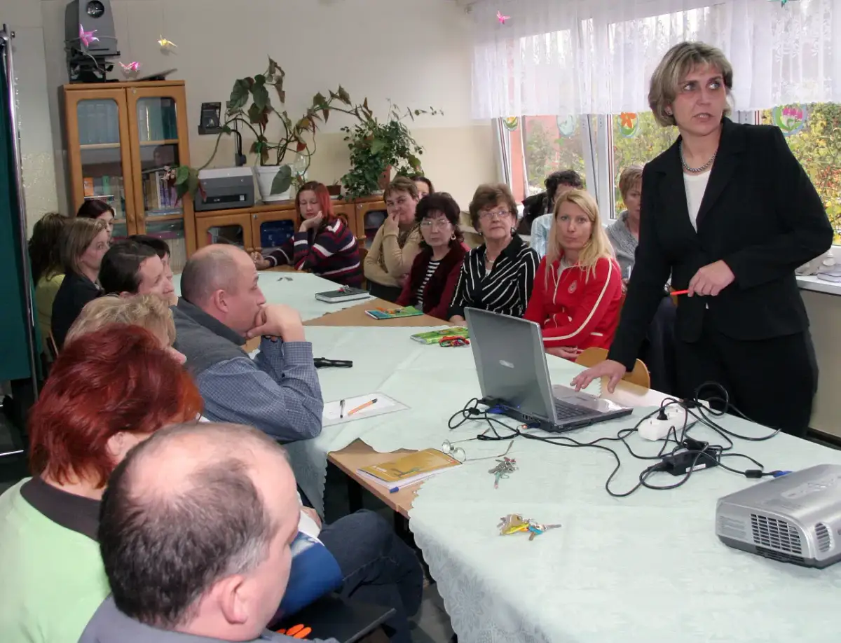 Nauczyciele zebrani na radzie pedagogicznej, dyskutują przy stole z laptopem i projektorem.