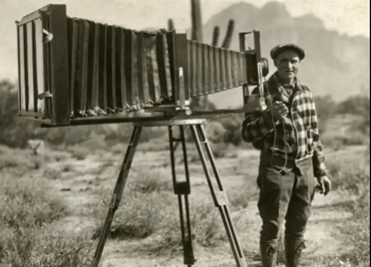 Mężczyzna w kratę stoi obok zabytkowego aparatu fotograficznego na statywie na pustynnym tle.