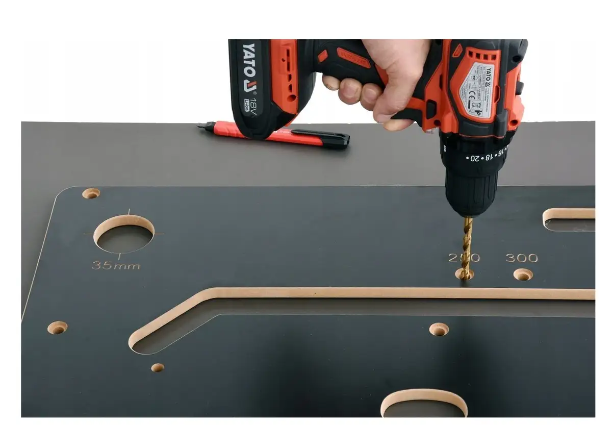 Dłoń trzyma wiertarkę, która wierci otwór w czarnym szablonie z oznaczeniami.