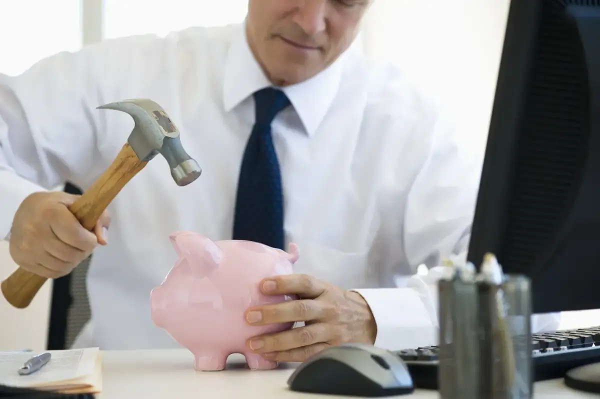 Mężczyzna w białej koszuli i krawacie zamierza rozbić skarbonkę młotkiem. Zastanawia się, ile trwa likwidacja lokaty, gdy pieniądze są w niebezpieczeństwie.