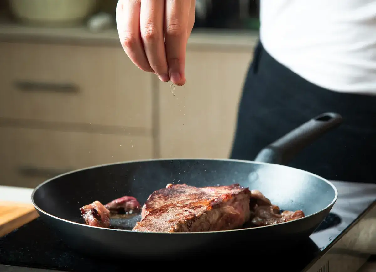 Ręka doprawia stek smażony na patelni.