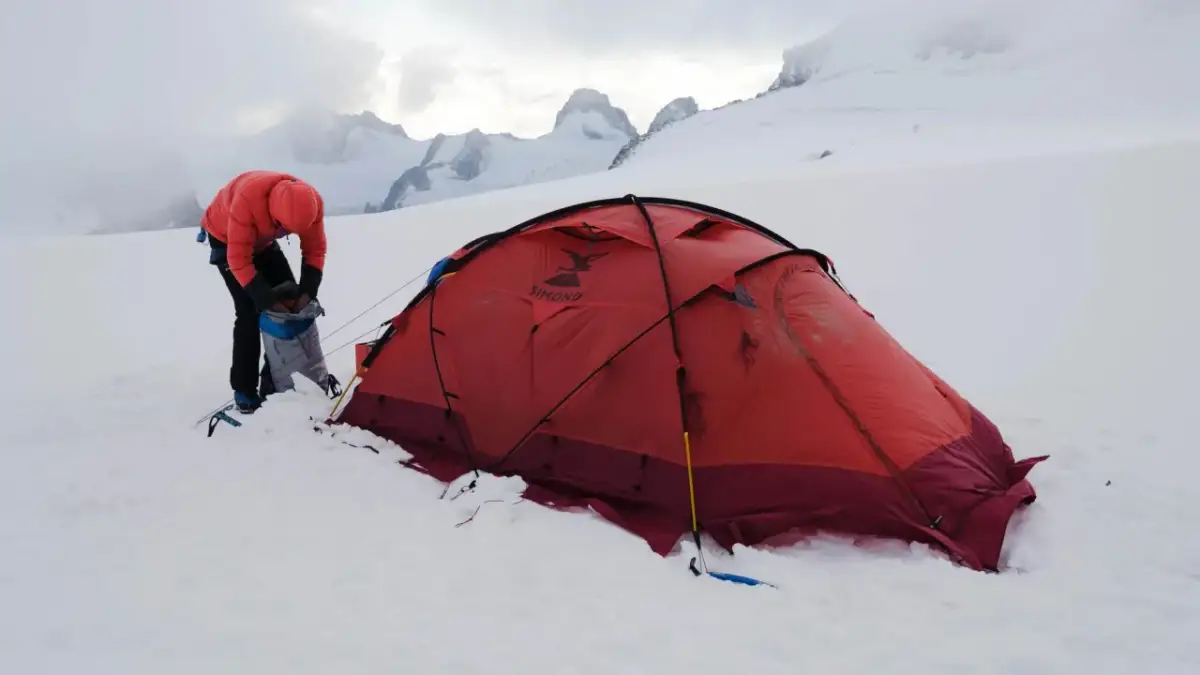 Osoba w pomarańczowej kurtce przygotowuje sprzęt przy czerwonym namiocie. W tle majestatyczne, ośnieżone szczyty, zapowiadające trudne wejście na Mount Everest.