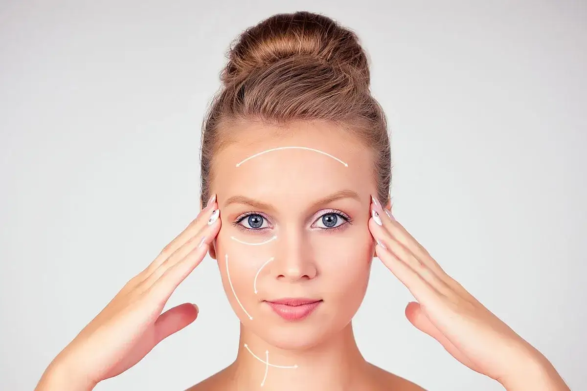 Młoda kobieta z włosami upiętymi w kok, z białymi strzałkami wskazującymi kierunek masażu twarzy.