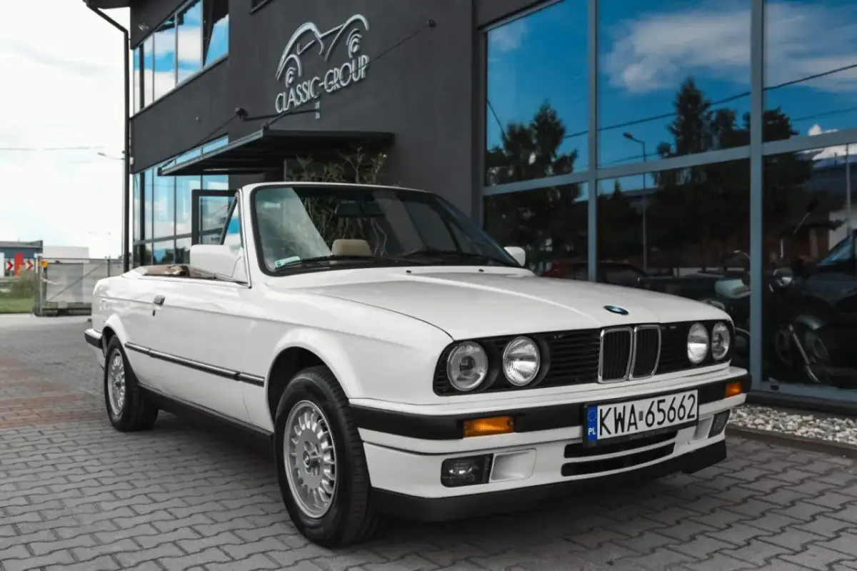 Białe bmw e30 cabrio z beżowym wnętrzem zaparkowane przed budynkiem z napisem "Classic Group".