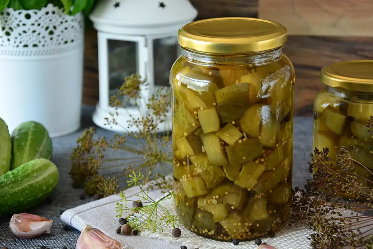 Słoiki pełne domowych pikli z ogórków, obok świeże ogórki, czosnek i koper.