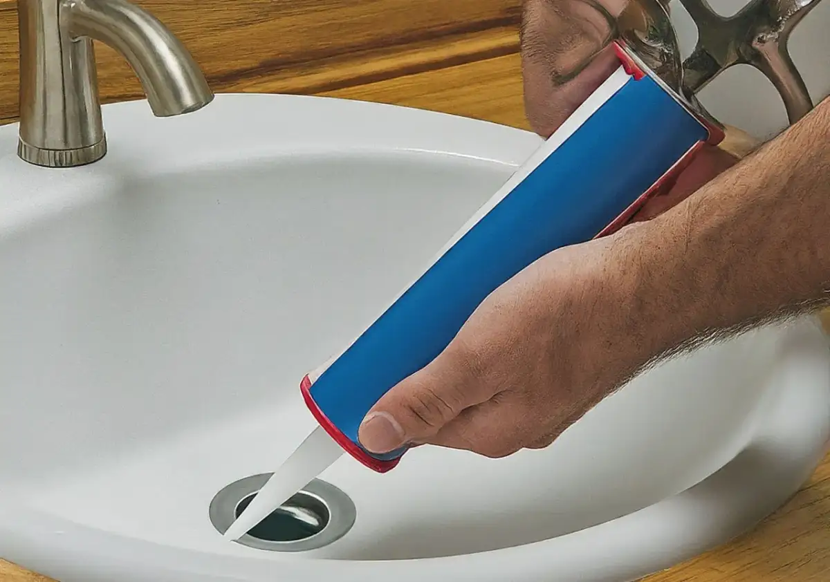 Dłoń trzymająca pistolet z silikonem, aplikując go wokół odpływu w białej umywalce. Czy silikon można nakładać warstwami?