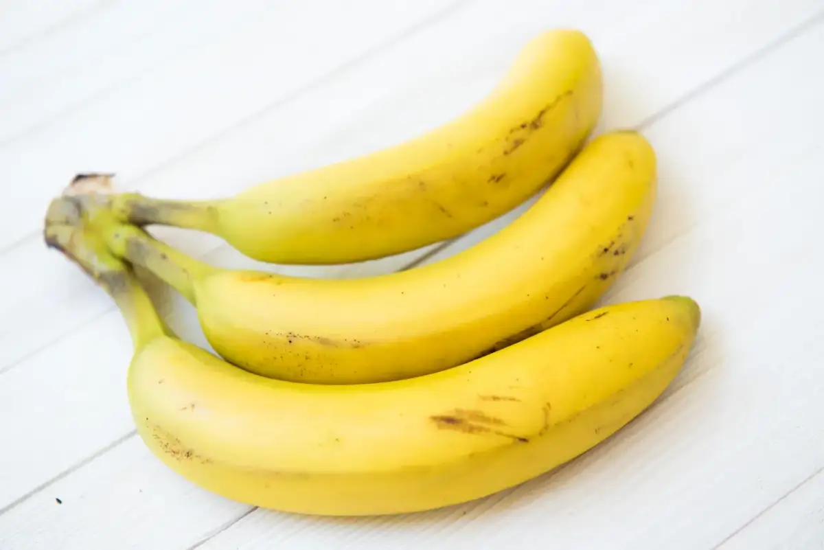 Trzy dojrzałe banany leżą na białym, drewnianym blacie. Czy banany tuczą? Zależy od ilości i sposobu ich spożycia.