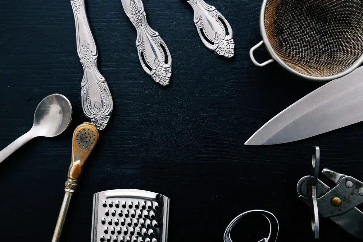Srebrne sztućce, sitko, nóż, tarka i nożyczki na czarnym tle.