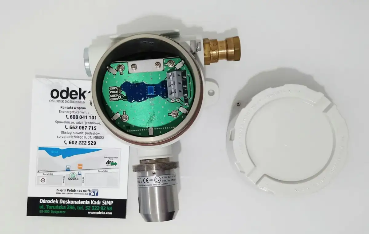 Otwarty detektor gazu z widoczną płytką drukowaną. Obok leży biała pokrywa z napisem "KEEP COVER TIGHT WHEN CIRCUITS ARE LIVE". To może być upg co to jest?
