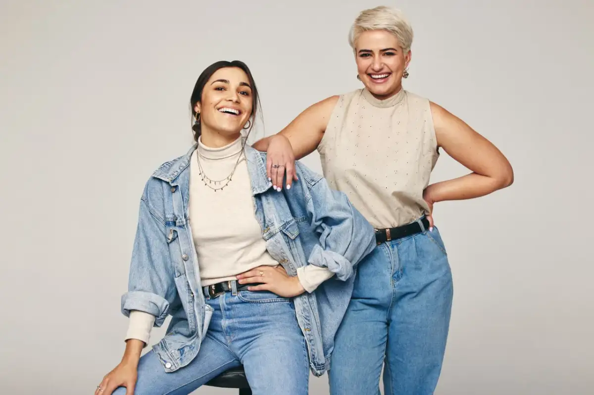 Dwie uśmiechnięte kobiety w jeansach. Jedna w beżowym golfie i jeansowej kurtce, druga w beżowej bluzce bez rękawów. Inspiracja: jaka bluzka do jeansów.
