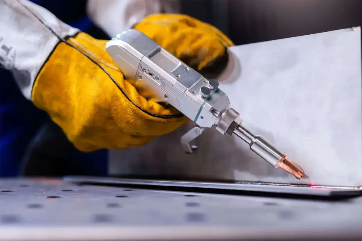 Ręce w żółtych rękawicach trzymają laserową spawarkę, która precyzyjnie łączy metalowe elementy, minimalizując wady spawalnicze.
