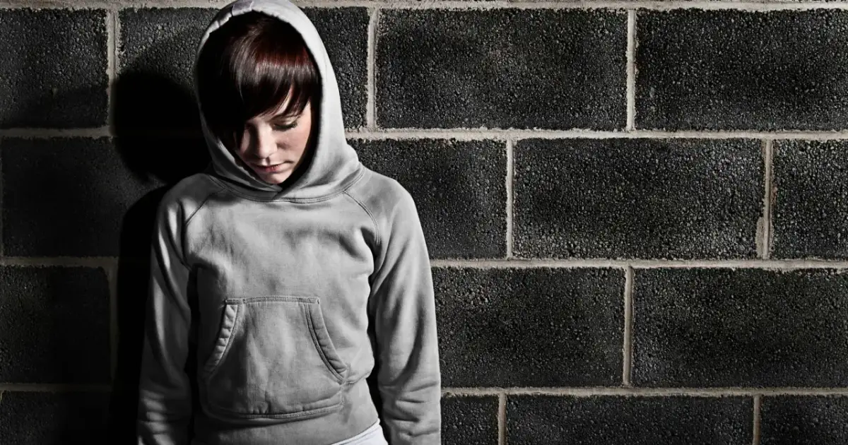 Smutna dziewczyna w szarej bluzie z kapturem stoi przy ceglanej ścianie. Może to być scena z poprawczaka dla dzieci, gdzie szuka nadziei.