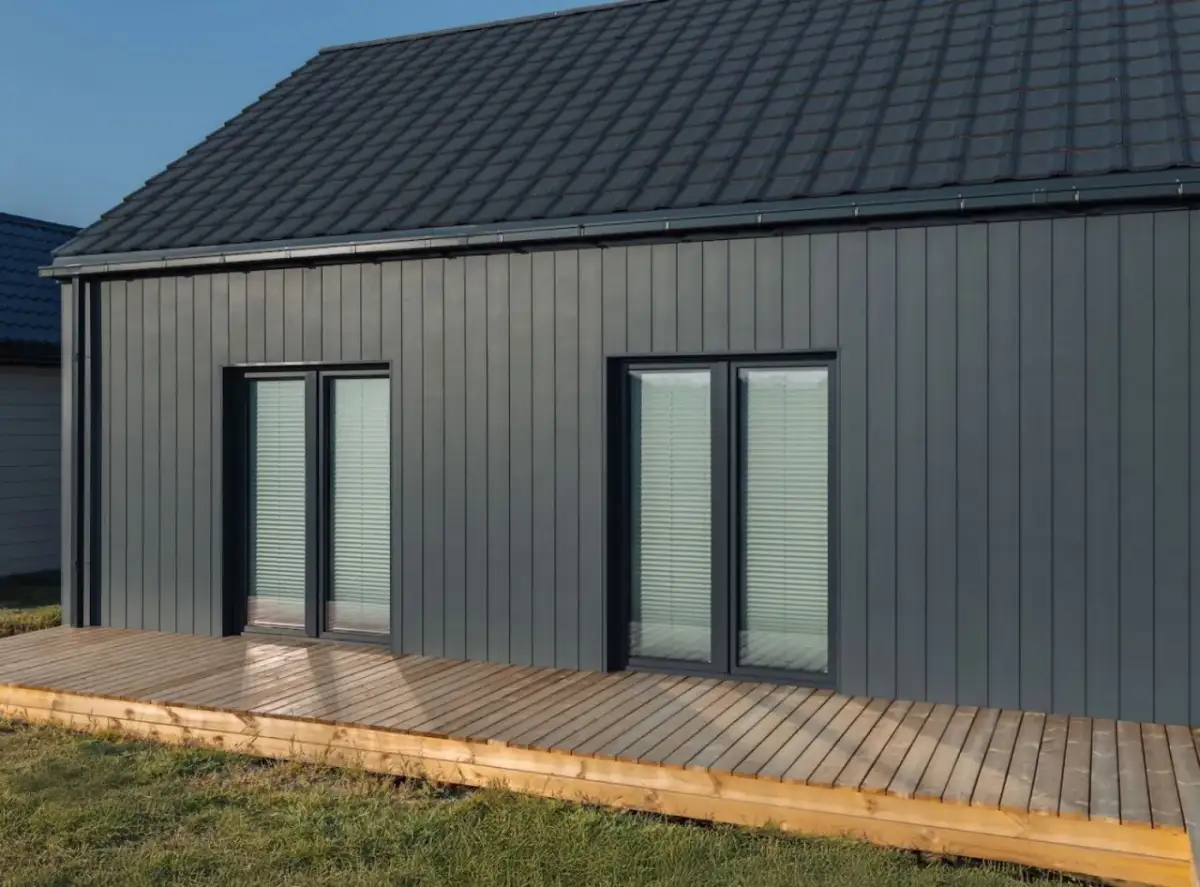 Nowoczesny dom z ciemnoszarą elewacją, co na styropian zamiast tynku? Drewniany taras i zieleń wokół.